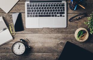 Workstation setup featuring a laptop, smartphone, clock, and stationery on a wooden desk during daytime photo