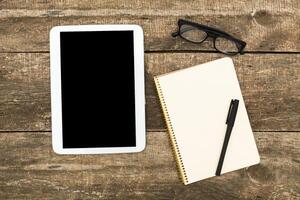 Workspace setup with tablet, notebook, pen, and glasses on rustic wooden table during daytime photo