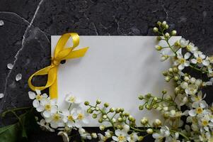 A flat-lay arrangement featuring a blank white card tied with a yellow ribbon, surrounded by delicate white flowers on a dark, textured surface photo