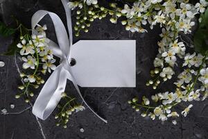 A blank white card is tied with a bright yellow ribbon on the upper left corner. White blossoms frame the bottom and right edges of the card, set against a dark gray, textured surface, photo
