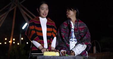 Handheld low angle view shot, Two Asian women dressed in warm cloth talk and share a happy moment together while grilling food and enjoying the cold night. video