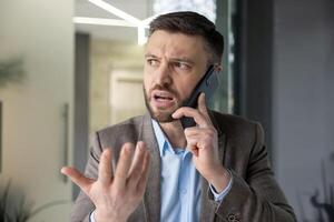A frustrated businessman in a suit is engaged in a heated phone conversation, expressing his emotions with a hand gesture in an office setting. photo