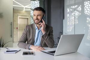 Smiling businessman talking on the phone while sitting at a desk in a modern office workspace with laptop and notepad. photo