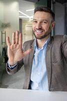 Smiling businessman waving hand at camera while making call in a modern office setting, showing cheerful greeting gesture photo
