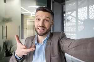 Businessman in a modern office making a call, smiling and engaging with the camera while gesturing photo