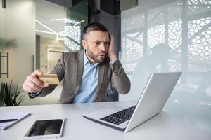 Man looking worried while shopping online with a credit card in a modern office, expressing concern about an online transaction photo