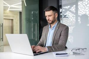 Professional businessman working on a laptop in a modern office setting, focused on his tasks. Background features glass walls and a notebook on the desk. photo