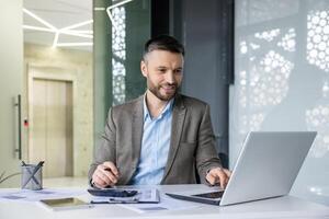 Smiling businessman working on laptop in modern office, analyzing documents and taking notes. Professional and focused in corporate environment. photo