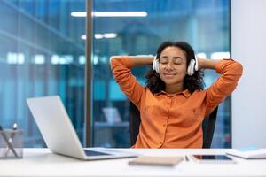 Office worker relaxing and resting listening to music on headphones while sitting at desk inside office. Businesswoman with arms folded behind head dreaming and thinking. photo