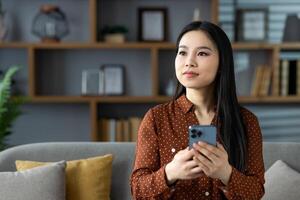 Asian woman with smartphone seated on sofa, gazing thoughtfully while surrounded by home comforts. Perfect for depicting technology, lifestyle, relaxation, and modern communication themes. photo