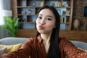 Asian woman taking selfie at home with playful expression. Captures modern lifestyle, technology use, and cozy atmosphere in living room setting, highlighting joy and self-expression. photo