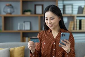 Asian woman using phone and card for online shopping at home. Relaxed on sofa, she engages in digital transactions, highlighting modern convenience and technology integration in daily life. photo