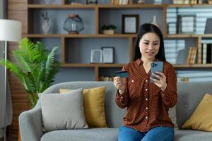 Asian woman at home, holding smartphone and credit card, smiling while engaging in online shopping. Comfortable environment, modern lifestyle, and convenience of digital transactions emphasized. photo