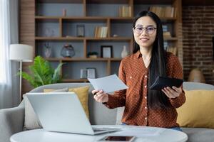Asian woman working from home, analyzing financial documents with laptop and calculator. Smiling, focused on task, showcasing multitasking and productivity in cozy, modern living room setting. photo