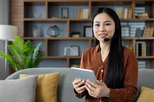 Asian woman with headset using tablet at home, smiling, seated on couch. Concept of remote work, online communication, technology. Ideal for virtual meeting, customer service, or e-learning. photo