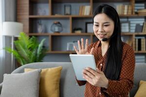 Asian woman participates in virtual meeting at home using tablet and headset. She is sitting on sofa, waving and smiling, demonstrating technology's role in remote communication. photo