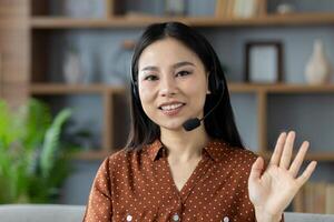 Cheerful Asian woman wearing headset, participating in online communication from comfortable setting, waves with friendly gesture. Image captures remote work, technology, connection, comfort, interaction. photo