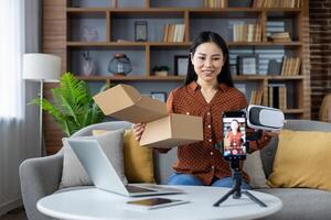 Asian woman sitting on sofa unboxing virtual reality headset during live stream. Smiling content creator using smartphone to engage audience. Concept of technology, communication, online business. photo