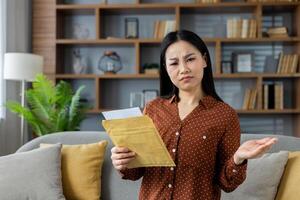 Asian woman with perplexed look holds envelope, expressing confusion over its contents. Scene captures emotion and curiosity in a cozy home environment, highlighting surprise. photo