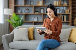 Asian woman with headphones happily engaged on tablet. Seated on sofa in cozy setting. Relaxed, modern home environment with natural elements. Technology use in comfortable space. photo