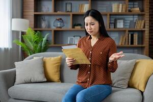 Asian woman sitting on sofa looks perplexed at letter, experiencing confusion or uncertainty. Home scene with bookshelf in background signifies indoor, personal setting. Emotional expression evident. photo