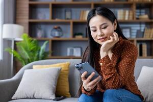 Asian woman in casual wear sitting on sofa using smartphone, smiling with relaxed expression. Modern home setting with decorative elements, enjoying leisure time indoors. photo