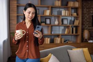 Asian woman holds smartphone and coffee, standing by cozy couch with bookshelf. Relaxed indoor setting, reading message on phone, enjoying leisure time at home. photo