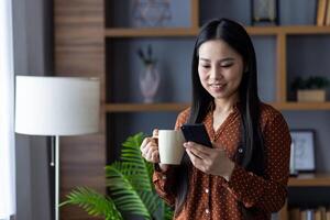 Asian woman at home smiling while using smartphone and drinking coffee. Casual and relaxed, she's in brown polka dot blouse enjoying modern communication in elegant home environment. photo