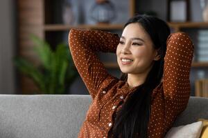 Asian woman in polka dot blouse enjoying relaxation at home, smiling as she leans back on sofa. Her peaceful expression conveys happiness and contentment in cozy setting. photo
