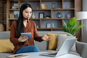 Asian woman using laptop and tablet for call. She wears headset, working remotely from home office. Concept includes remote work, virtual communication, modern technology. photo