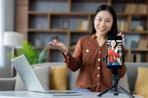 Asian woman using smartphone on tripod at home, recording vlog. Smiling, engaging, laptop visible, sharing ideas. Perfect for illustrating digital content creation, at-home productivity. photo