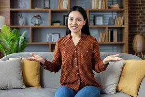 Cheerful asian woman sitting on cozy sofa sharing friendly conversation at home. Relaxed atmosphere with plants, books all around. Positive expression, open gesture, casual clothing. photo