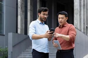 Two serious colleagues are chatting and talking outside the office building. Men in casual clothes are discussing new app on smartphone, online chart, investment report. photo