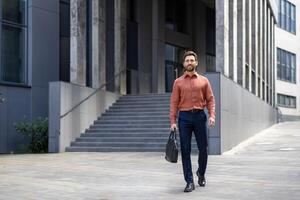 A successful businessman in full growth walks through the city with a business bag in his hands, a mature man smiles contentedly from outside an office building. photo
