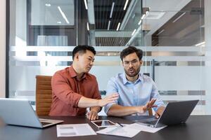 Two colleagues in casual clothes discussing a work project, men sitting at a desk with a laptop. Viewing electronic online financial reports. photo