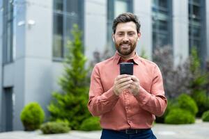 Successful man walking through the city outside an office building with a phone in his hands. Businessman using an app on his smartphone, typing messages, and browsing social media, smiling. photo