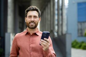 Portrait of successful businessman man with phone in hands. Office worker smiling and looking at camera, using app on smartphone, walking in city from outside office building. photo