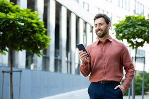 Successful man walking through the city outside an office building with a phone in his hands. Businessman using an app on his smartphone, typing messages, and browsing social media, smiling. photo