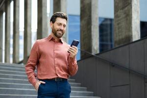 Successful man walking through the city outside an office building with a phone in his hands. Businessman using an app on his smartphone, typing messages, and browsing social media, smiling. photo