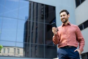 Successful man using an app on his smartphone, businessman walking around the city smiling happily typing a text message from outside an office building. photo