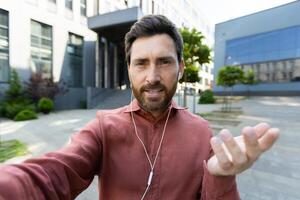 Man with headphones looking into phone camera. Office worker businessman talking to colleagues and friends walking around the city using call, smartphone app. photo