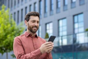 Businessman smiling as he uses smartphone outside modern office building. Casual attire and positive expression suggest satisfaction, connectivity, engagement in digital communication photo