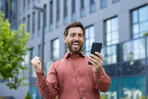 A man is holding a cell phone and smiling. He is standing in front of a building. photo