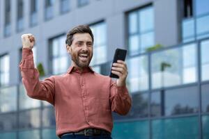 A man is holding a cell phone and smiling. He is wearing a red shirt. The building behind him is gray photo