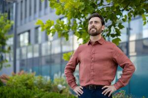 A man in a red shirt stands in front of a building. He is smiling and looking up at the sky photo