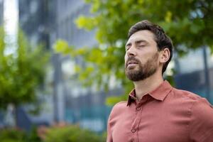 Mature male professional standing outside in city with eyes closed, experiencing calm and relaxation. Businessman taking break, capturing serene and positive emotions in urban setting a photo