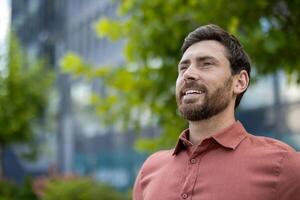 A man with a beard and a red shirt is smiling. He is standing in front of a building photo