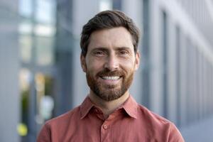 A man with a beard and a red shirt is smiling. He is standing in front of a building photo