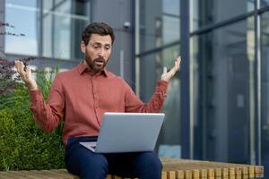 Mature man sitting outdoors in urban setting, using laptop with surprised expression. Wearing casual attire, he reacts to unexpected online news or communication, illustrating shock and emotion. photo