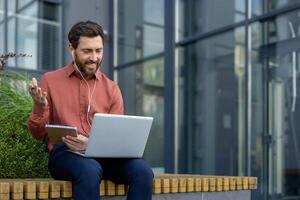 Mature man on call with laptop and headphones, sitting outdoors. He appears engaged and focused while gesturing and holding notebook, reflecting business communication and remote work concepts. photo